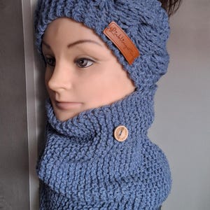 Può includere: Un set di cappello e scaldacollo in maglia blu. Il cappello ha un'apertura per la coda di cavallo e un'etichetta in pelle marrone con testo. Lo scaldacollo ha una chiusura con bottone in legno. Il set è esposto su una testa di manichino.
