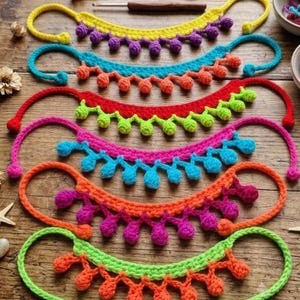 Puede incluir: Seis guirnaldas de ganchillo en forma de media luna dispuestas verticalmente, cada una con una combinación de colores diferente de hilo y pompones colgantes. Los colores incluyen amarillo, turquesa, rojo, rosa, naranja y verde lima.
