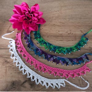 May include: Four crocheted necklaces in white, pink, purple-blue, and green-blue gradients, each featuring a pointed lace-like edge accented with small silver beads. A large, vibrant fuchsia dahlia flower is positioned at the top left corner.