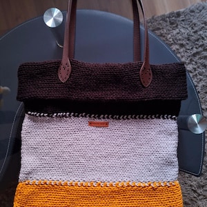Könnte beinhalten: Handgefertigte Tragetasche mit braunen Ledergriffen und -akzenten. Die Tasche hat ein Strickdesign in Braun-, Grau- und Orangetönen. Ein kleines rechteckiges Etikett mit der Aufschrift "handmade" ist angebracht. Der Text "sk.headbands" ist sichtbar.