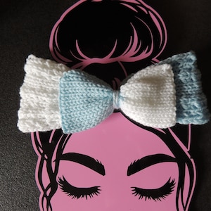 Könnte beinhalten: Eine weiße und hellblaue gestrickte Schleife mit einer mittleren Schleife. Die Schleife befindet sich auf einem rosa Hintergrund mit einer Silhouette eines Frauenkopfes.