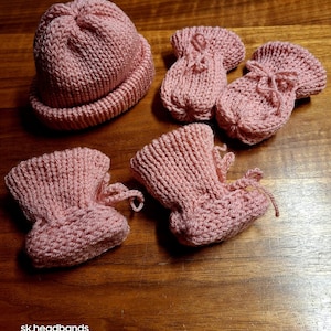 Könnte beinhalten: Ein gestricktes Baby-Set in zartrosa, bestehend aus einer Mütze und drei Paar Babyschuhen. Die Babyschuhe haben einen Kordelzugverschluss. Der Text "sk.headbands" ist unten sichtbar.