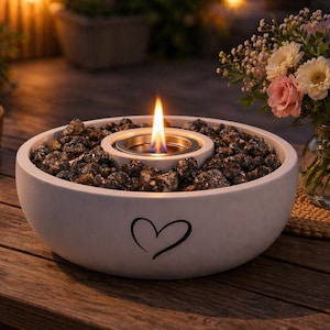 Könnte beinhalten: Eine weiße Feuerschale mit Herzmotiv, gefüllt mit dunklen Steinen und einer Flamme. Auf einem Holztisch daneben steht ein Blumenstrauß in einer Vase. Ein kleiner Korb und ein gefaltetes Tuch ergänzen die Szene.