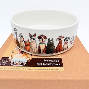 Könnte beinhalten: Eine weiße Keramik-Hundenapf mit einer bunten Cartoon-Illustration verschiedener Hunderassen. Die Schüssel steht auf einem braunen Karton. Auf der Box steht "Für Hunde mit Geschmack."