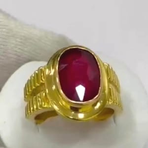 Puede incluir: Un anillo de oro con una gran gema de rubí de corte ovalado. El anillo tiene un diseño detallado y texturizado en la banda y alrededor del engaste de la gema. El rubí es de color rojo intenso.