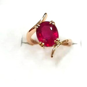 以下が含まれることがあります： 大きなオーバルカットのルビーをあしらった、ローズゴールドのリング。ルビーは鮮やかな赤色で、繊細な爪で固定されています。リングのバンドは、ユニークで流れるようなデザインです。
