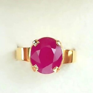 May include: A gold ring with a large, oval-cut ruby gemstone. The ruby is a vibrant red colour, and the ring's band is polished gold. The gemstone is held in place by four gold prongs.