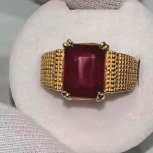 Peut inclure: Une bague en or avec une grosse pierre précieuse rouge foncé de forme rectangulaire. La bague a une bande texturée et des accents dorés décoratifs autour de la pierre précieuse. La bague est présentée sur une surface blanche.