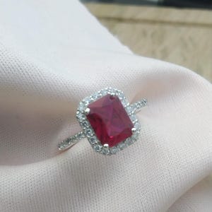 May include: A silver ring with a large, rectangular, ruby-red gemstone, encircled by a halo of smaller, clear stones. The ring's band is also set with small, clear stones, creating a sparkling effect. The ring is displayed on a soft, pink fabric.