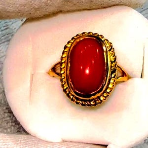 May include: A gold ring featuring a large, oval-shaped, polished red gemstone. The gemstone is set in a gold frame with a rope-like design. The ring is presented on a white surface, suggesting a jewelry display.