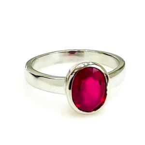 May include: A silver ring featuring a large, oval-cut, vibrant red gemstone. The band is smooth and polished, with a simple, elegant design. The gemstone is set in a bezel setting, enhancing its color and brilliance.