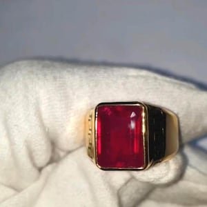 May include: A gold ring featuring a large, rectangular, deep red gemstone. The ring has a classic design with a gold band and black detailing. The gemstone is the focal point, catching the light and adding a touch of luxury and style.