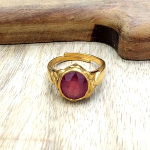以下が含まれることがあります： 大きな楕円形の赤い宝石が付いた、金色の指輪。 指輪には調節可能なバンドがあり、宝石のセッティングの側面に装飾的なディテールがあります。 指輪は明るい色の木製の表面に表示されています。