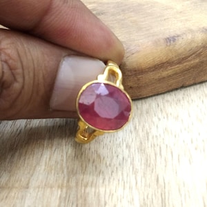 以下が含まれることがあります： 大きなオーバルカットのルビーの宝石をあしらったゴールドリング。ルビーは深い赤色で、ゴールドのベゼルにセットされています。リングはシンプルでエレガントなデザインです。