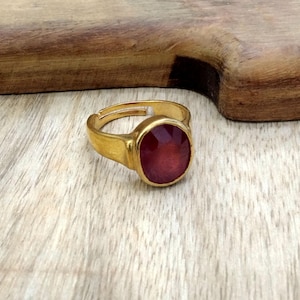 Op de afbeelding: Een goudkleurige verstelbare ring met een grote, ovaalvormige, gefacetteerde rode edelsteen. De ring ligt op een lichtgekleurd houten oppervlak. De edelsteen heeft een dieprode kleur en reflecteert licht.