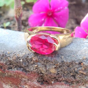 Puede incluir: Un anillo de oro con una gran gema roja rubí de forma ovalada. El anillo está colocado sobre una superficie de piedra gris rugosa, con flores rosas en el fondo borroso. La banda del anillo es gruesa y pulida.