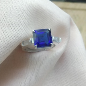 Könnte beinhalten: Ein silberner Ring mit einem großen, quadratisch geschliffenen, tiefblauen Edelstein als Mittelpunkt. Zwei kleinere, rechteckige Steine flankieren den Hauptstein. Der Ring ist vor einem weichen, hellen Hintergrund platziert.