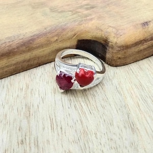 May include: A silver ring featuring two heart-shaped gemstones, one deep red and the other a vibrant red. The ring has a double band design with small, clear accent stones. The ring is displayed on a light wood surface.