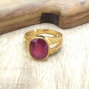 以下が含まれることがあります： オーバルカットのルビー色の石が付いた、金色の指輪。 指輪には、装飾的なデザインのテクスチャード加工されたバンドが付いています。 明るい色の木製の表面に置かれています。