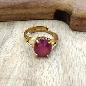以下が含まれることがあります： 大きな楕円形の深い赤色の宝石をあしらった、ゴールドカラーの調節可能なリング。リングのバンドは、宝石の両側に装飾的な要素があり、明るい木材の表面にセットされたオープンデザインです。