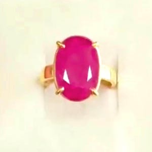 May include: A gold ring with a large, oval-shaped, bright pink gemstone. The gemstone is secured by four gold prongs, and the band is plain, polished gold. The ring is displayed against a plain background.