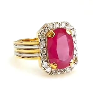 Könnte beinhalten: Ein goldener Ring mit einem großen, oval geschliffenen Rubin als Mittelstein. Der Rubin ist von einem Kranz aus kleinen, klaren Diamanten umgeben. Das Ringband hat ein geschichtetes Design aus Gold und Silber.