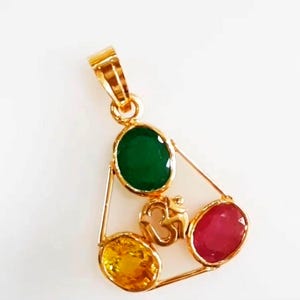 May include: A gold pendant featuring three oval gemstones: green, yellow, and red. The gemstones are set within a gold frame, with a gold Om symbol in the center. The pendant has a gold loop for a chain.