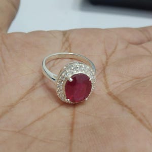 Peut inclure: Une bague en argent avec une grosse pierre centrale en rubis de forme ovale. Le rubis est entouré d'un halo de petites pierres précieuses claires. La bague est montée sur un anneau en argent, présentant un design classique et élégant.