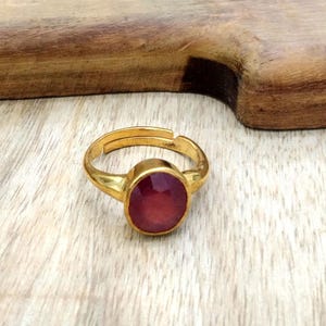 以下が含まれることがあります： 大きな楕円形の赤い宝石が付いた、金色の指輪。明るい色の木製の表面に置かれています。宝石は焦点であり、研磨された反射面を持っています。