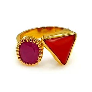 Könnte beinhalten: Ein goldfarbener Ring mit zwei markanten Edelsteinen. Einer ist ein runder, magentafarbener Stein mit einer goldfarbenen, gewellten Fassung. Der andere ist ein dreieckiger, leuchtend roter Stein. Der Ring hat ein modernes, geometrisches Design.