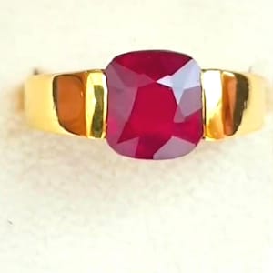May include: A gold ring with a large, cushion-cut ruby gemstone. The ruby is a deep red colour and is set in a gold band. The ring has a classic, elegant design.