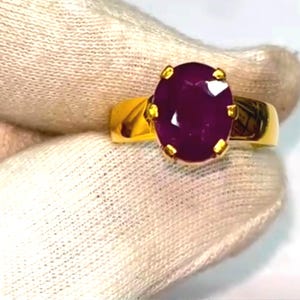 May include: A gold ring with a large, oval-cut, deep red gemstone. The gemstone is secured by four gold prongs. The ring's band is a simple, polished gold design. The ring is shown against a white background.