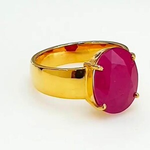 May include: A gold ring featuring a large, oval magenta gemstone. The ring has a wide, polished band and a setting that secures the gemstone. The ring is set against a white background.