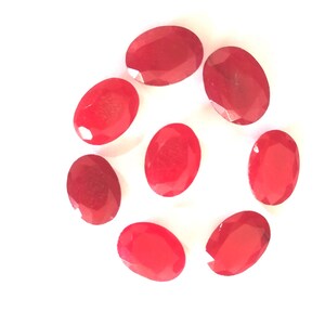 Può includere: Otto gemme ovali sfaccettate di un vivace colore rosso sono disposte in un motivo circolare su uno sfondo bianco. Le gemme hanno un aspetto lucido e brillante, che riflette la luce.