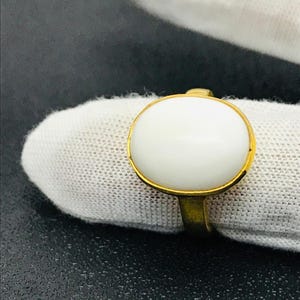 May include: A gold ring with a large, oval, white stone. The stone is set in a gold bezel, and the band has a simple design. The ring is shown on white fabric. A classic piece of jewellery.