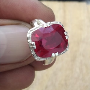 May include: A silver ring featuring a large, cushion-cut, deep red gemstone. The gemstone is set in a detailed, ornate silver band. The ring is held up against a blurred background, showcasing the vibrant color of the stone.