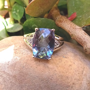 Op de afbeelding: Een zilveren ring met een grote, rechthoekige, blauwe edelsteen. De edelsteen heeft meerdere facetten die het licht reflecteren. De ring heeft kleinere accentstenen aan weerszijden van de hoofdsteen. De ring ligt op een natuurstenen oppervlak.