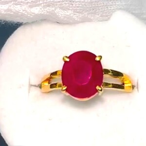 以下が含まれることがあります： 大きな楕円形のルビーの宝石をあしらったゴールドリング。ルビーは深い赤色で、スプリットシャンクのデザインのゴールドバンドにセットされています。リングは白い表面に表示されています。