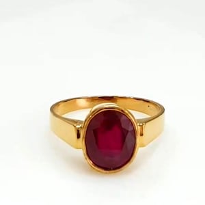 May include: A gold ring with a large, oval-shaped, deep red gemstone. The ring has a simple, classic design with a polished gold band. The gemstone is the focal point, set in a bezel setting, highlighting its rich colour and brilliance.