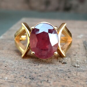 以下が含まれることがあります： 大きな楕円形のルビーの宝石をあしらったゴールドリング。ルビーは深い赤色で、側面にユニークな幾何学模様が施されたゴールドバンドにセットされています。リングは質感のある表面に表示されています。