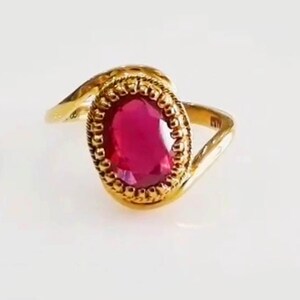 May include: A gold ring with an oval, deep red gemstone. The gemstone is framed by a detailed, beaded border. The ring's band has a curved design, set against a white background.