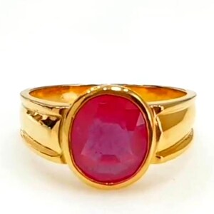 May include: A gold ring with a large, oval ruby gemstone. The ring has a wide, polished band. The ruby is set in a bezel setting. The ring is shown against a white background. The ring is a piece of jewellery.