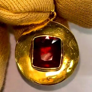Könnte beinhalten: Ein goldener Anhänger mit einem großen, quadratisch geschliffenen, tiefroten Edelstein in der Mitte. Der Anhänger hat einen kreisförmigen, strukturierten Goldrahmen und eine kleine Schlaufe für eine Kette. Der Edelstein ist facettiert.