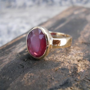以下が含まれることがあります： 大きなオーバルカットのルビーの宝石が付いたゴールドリング。 ルビーは深い赤色で、磨かれたゴールドバンドにセットされています。 リングは風化した木製の表面に表示されています。