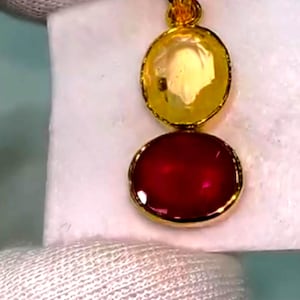May include: A gold pendant featuring two gemstones. The top stone is a round, yellow gem, and the bottom is an oval, deep red gem. The pendant is suspended from a gold chain loop, showcasing a classic jewelry design.