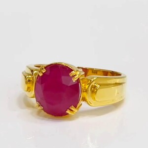 Op de afbeelding: Een gouden ring met een grote, ovaal geslepen robijn. De robijn heeft een dieprode kleur en is gezet in een gouden band met een klassiek ontwerp. De ring wordt gepresenteerd op een wit oppervlak.