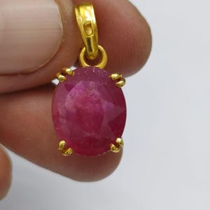 May include: A gold pendant with a large, oval-cut ruby gemstone. The ruby is a deep red colour and is held by four gold prongs. The pendant has a simple, elegant design, suitable for everyday wear.