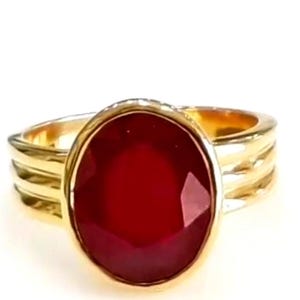 May include: A gold ring with a large, oval-cut, deep red gemstone. The ring has a textured band with three parallel ridges. The gemstone is the focal point, set in a gold bezel.