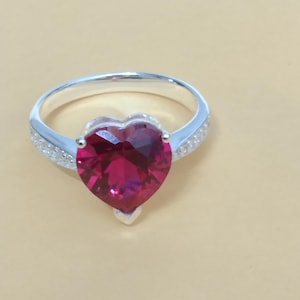 Peut inclure: Une bague en argent ornée d'une grosse pierre précieuse rouge foncé en forme de cœur. Le groupe est en argent avec de petites pierres d'accent claires. La pierre précieuse est facettée, captant la lumière et créant un effet scintillant.