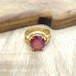 以下が含まれることがあります： 大きな楕円形の赤い宝石が付いた、金色の指輪。 指輪には調節可能なバンドがあり、宝石のセッティングの側面に装飾的なアクセントがあります。 指輪は明るい色の木製の表面に表示されています。